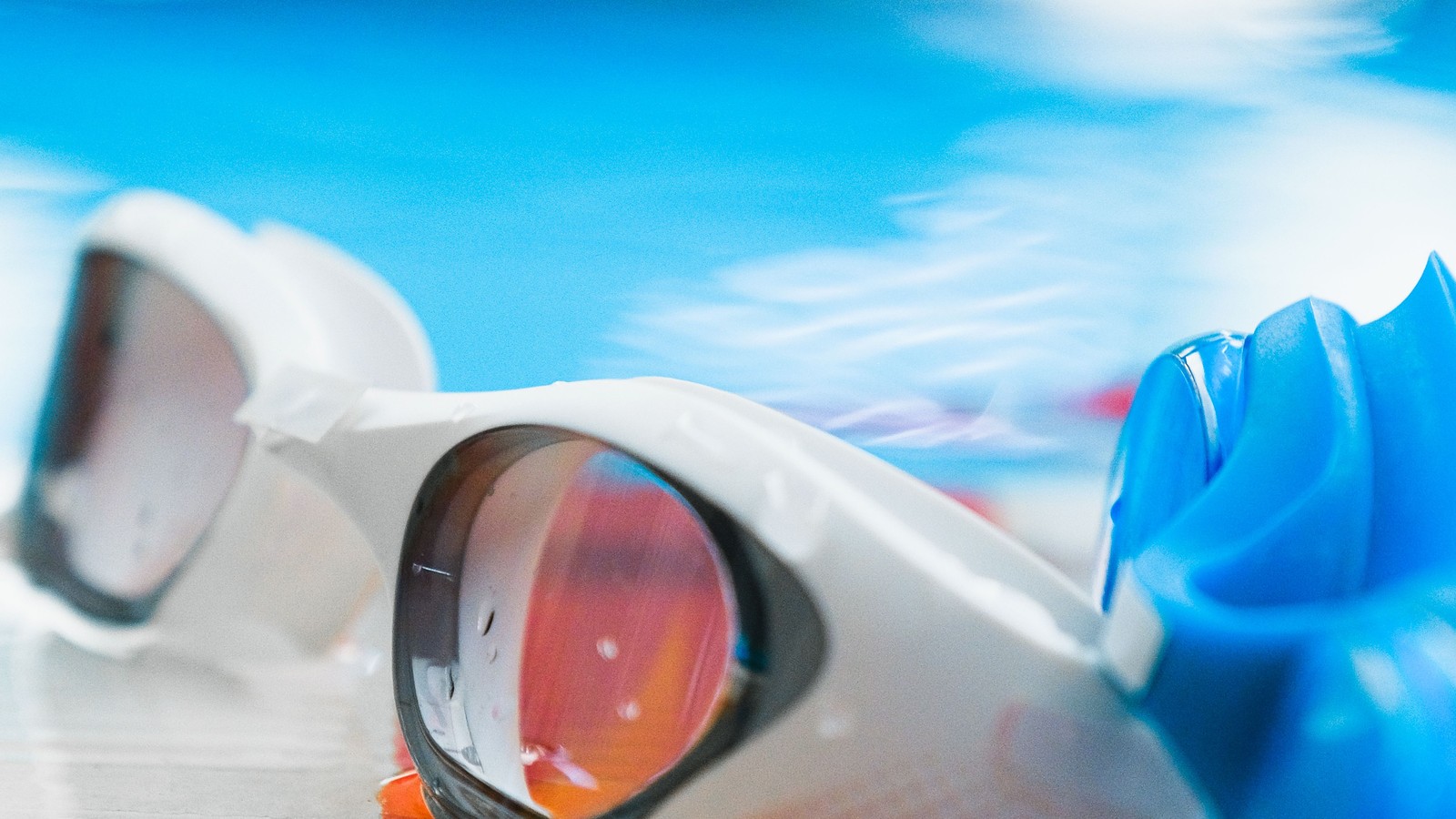 White swim goggles and a blue swim cap resting on a wet poolside surface.